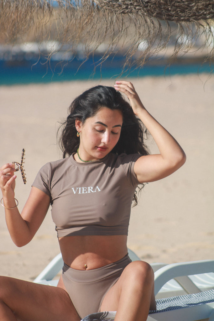 Woman in a brown 'Viera' swimsuit sitting on a beach chair by the ocean.